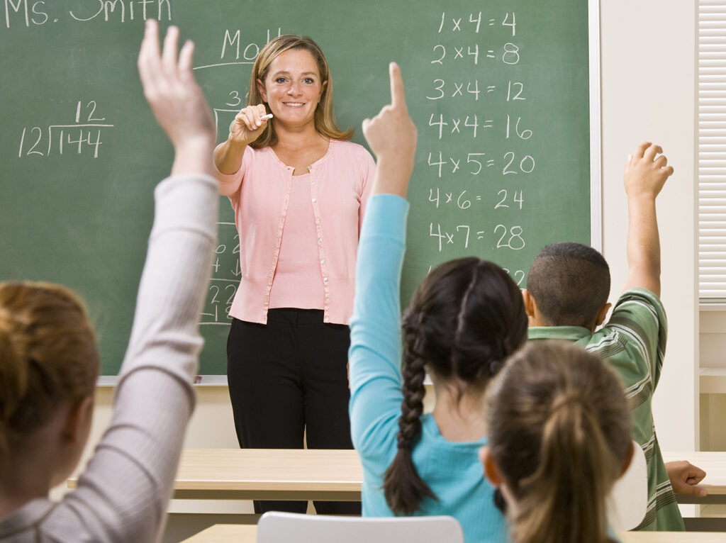 Nauczycielka, za nią tablica, przed nią dzieci zgłaszające się do odpowiedzi