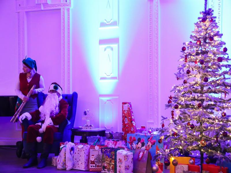 Mikołaj z Teatrem Domino w SCK - Zameczek. Mikołaj i elf siedzą przy choince, obok mnóstwo prezentów dla dzieci