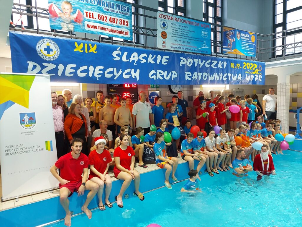 Uczestnicy zawodów stojący w grupie na brzegu basenu pod banerem.