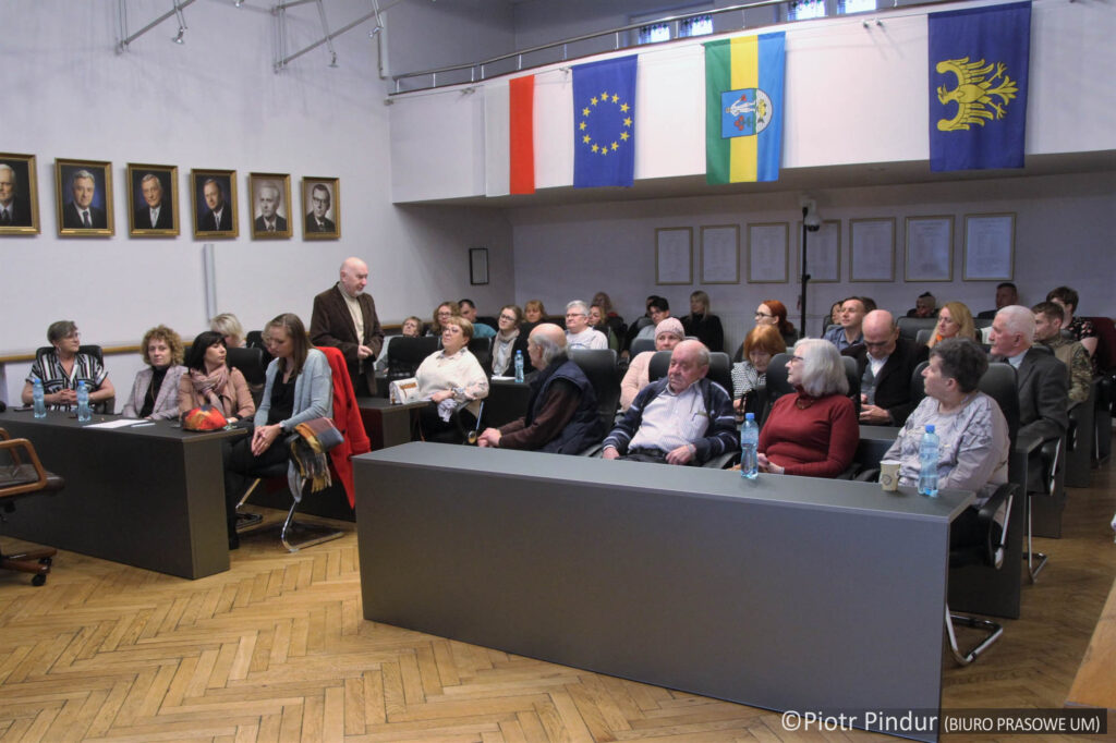 Kobiety i mężczyźni siedzący na krzesłach przy stołach w dużej sali. Po lewej stronie wiszące na ścianie portrety, po prawej, u góry wiszą flagi: Polski, Unii Europejskiej, Siemianowic Śląskich i Województwa Śląskiego