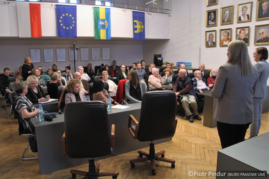 Na pierwszym planie dwie stojące kobiety, tyłem do obiektywu. Na drugim planie kobiety i mężczyźni siedzący w fotelach w dużej sali