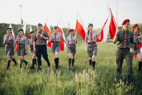 Harcerze i harcerki na łące z biało-czerwonymi flagami.