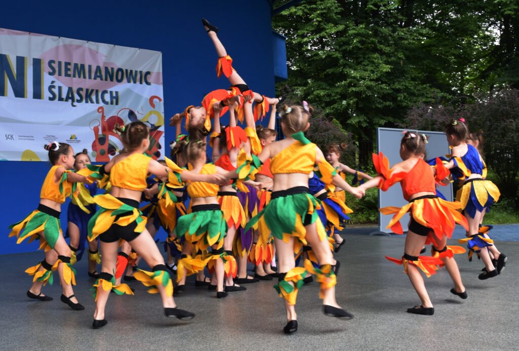 Tańczące w kole dzieci w kolorowych kostiumach i w środku wysoko podniesiona mała tancerka