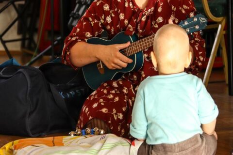 Zdjęcie zrobione podczas zajęć Maluszkowego Muzykowania w SCK - Bytków. Siedzące tyłem do obiektywu małe dziecko słucha grającej na gitarze prowadzącej zajęcia
