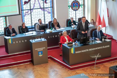 mężczyzna stoi za stołem prezydialnym i przemawia. Z tyłu przy innym stole prezydialnym siedzą kobiety i mężczyźni