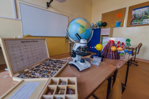 Pracownia szkolna, w tle tablica, na ławce szkolnej stoją pomoce naukowe: mikroskop, globus, minerały