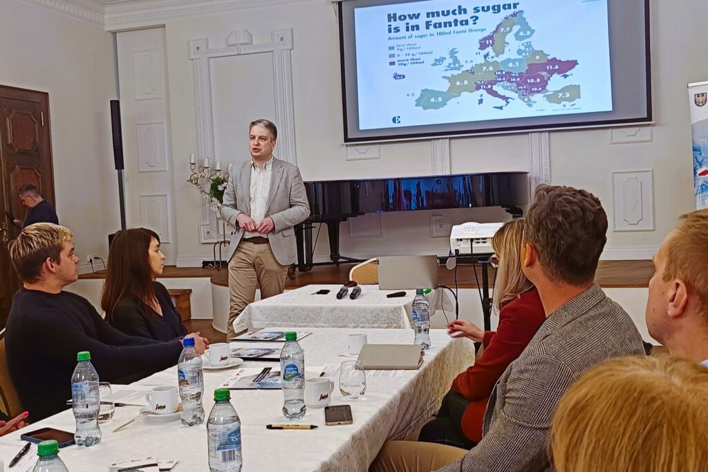 Mężczyzna w jasnej marynarce prowadzi prezentację dla uczestników spotkania. Na ekranie wyświetlona jest mapa Europy z porównaniem zawartości cukru w napoju Fanta.