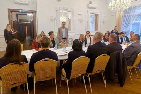 Uczestnicy spotkania, na środku mężczyzna w jasnej marynarce stoi i zabiera głos, pozostali siedzą wokół stołu.