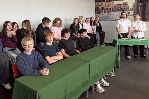 W sali lekcyjnej uczniowie siedzą w dwóch rzędach przy zielonych stołach, a naprzeciw nich stoją prowadzący w białych koszulach. W tle widoczna fototapeta z polską flagą i zabytkowymi kamienicami Nikiszowca.
