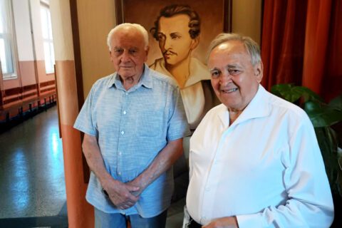 Dwaj starsi mężczyźni – Zbigniew Paweł Szandar oraz prof. Józef Śliwiok – stoją obok siebie w korytarzu Szkoły Podstawowej nr 11 w Siemianowicach Śląskich. Za nimi widać portret Juliusza Słowackiego.