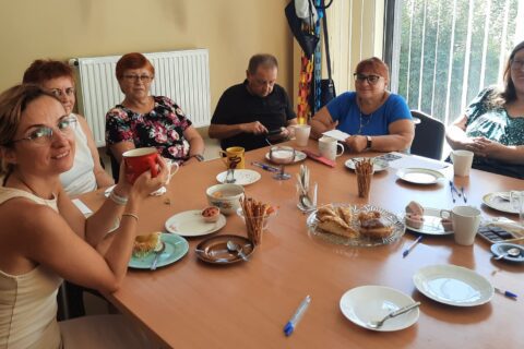 Grupa seniorów i osób z niepełnosprawnościami siedzi przy wspólnym stole w sali zajęciowej. Uczestnicy rozmawiają, część się uśmiecha, na stole leżą kubki i notatki.