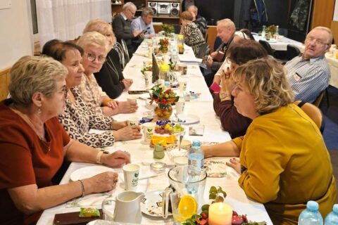 Wieloosobowa grupa siedzi przy wspólnym, zastawionym stole w sali z trofeami i tablicami informacyjnymi, na stole świeca, woda, przekąski i dekoracje.