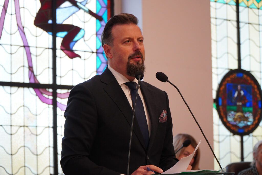 Prezydent miasta przemawia do radnych przy mównicy na tle witraży.