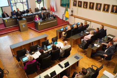 Sala obrad Rady Miasta wypełniona radnymi i urzędnikami, w tle flagi, witraże i portrety byłych włodarzy miasta.