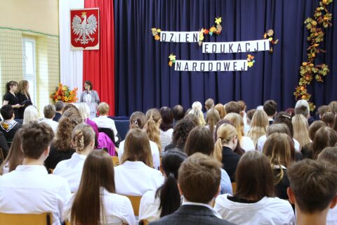Widok na salę z tłumem uczniów i nauczycieli słuchających przemówienia na tle polskich barw i orła oraz napisu „Dzień Edukacji Narodowej”