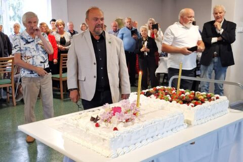 Prezes Jan Maj w beżowej marynarce stoi przy dużym białym torcie jubileuszowym w kształcie cyfry dziewięćdziesiąt, udekorowanym truskawkami i owocami. Wokół niego zebrani goście uroczystości, część trzyma telefony, by uwiecznić ten moment. W tle jasne ściany sali oraz obrazy na ścianach.