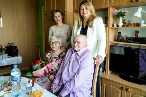 Wspólne zdjęcie uczestników spotkania. Siedząc: Pani Maria w kolorowej sukience i Pan Jan w fioletowej koszuli. Stojąc: Druga Zastępca Prezydenta Miasta Beata Gałecka, Kierownik USC Magdalena Mietły i Grzegorz Kukla. Na stole: bukiet róż, pamiątkowy dokument, zastawę, napoje, ciastka.