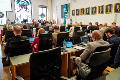 Sala sesyjna w czasie obrad. Radni siedzą w kilku rzędach, korzystając z laptopów i przeglądając dokumenty. W tle widać stół prezydialny oraz barwne witraże, które tworzą charakterystyczny klimat sali.
