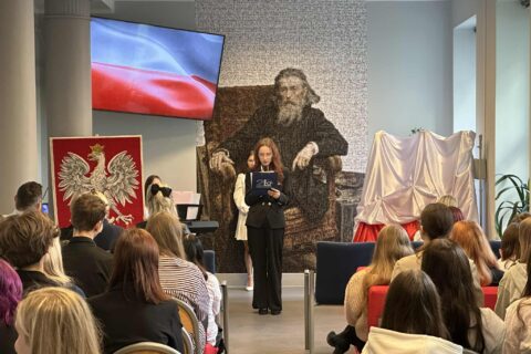 Dwie uczennice stoją przy mikrofonie podczas akademii w sali z polską flagą na ekranie i godłem Polski.