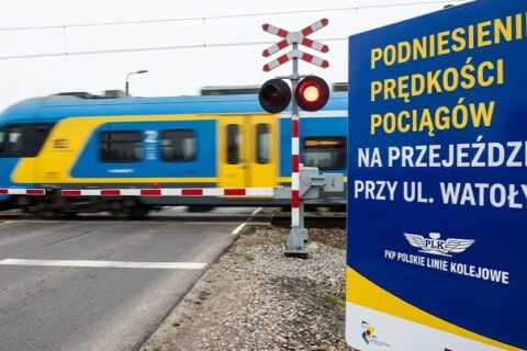 Zdjęcie przedstawia aktywny przejazd kolejowy. W tle widać szybko przejeżdżający, rozmyty pociąg pasażerski w barwach niebiesko-żółtych. Biało-czerwone rogatki są opuszczone, a na sygnalizatorze migają czerwone światła ostrzegawcze. Na pierwszym planie, po prawej stronie, stoi duża, ciemnoniebieska tablica informacyjna. Znajduje się na niej duży żółty i biały napis: „PODNIESIENIE PRĘDKOŚCI POCIĄGÓW NA PRZEJEŹDZIE PRZY UL. WATOŁY…”, a poniżej białe logo PKP Polskie Linie Kolejowe.