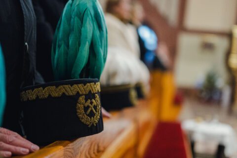 Zbliżenie na górniczą czapkę z zielonym pióropuszem, leżącą na ławce w kościele podczas mszy świętej. W tle rozmazane sylwetki uczestników uroczystości tworzą ciepłą, podniosłą atmosferę.