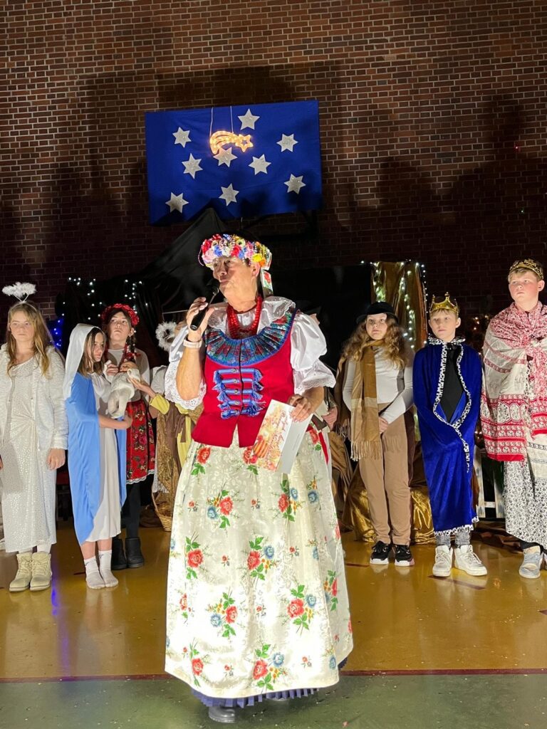 Na środku sali gimnastycznej stoi pani dyrektor w tradycyjnym śląskim stroju ludowym: białej bluzce, czerwonym gorsetem i długiej, wzorzystej spódnicy. W ręku trzyma mikrofon i kartkę z życzeniami. Za nią w półokręgu stoją dzieci przebrane za postacie z jasełek, m.in. aniołów, pasterzy i królów. W tle widoczna jest świąteczna scenografia ze gwiazdami i oświetleniem.