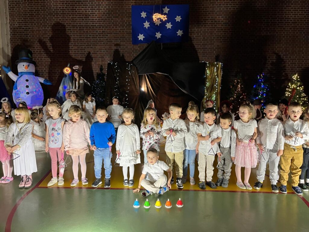 Starsze przedszkolaki siedzą i stoją na scenie, zwrócone w stronę prowadzącej nauczycielki, która klęczy na pierwszym planie i dyryguje. Dzieci ubrane są w jasne stroje i uważnie obserwują jej gesty. Za nimi widoczna jest bogata bożonarodzeniowa dekoracja z choinkami, światłami i postaciami jasełkowymi.