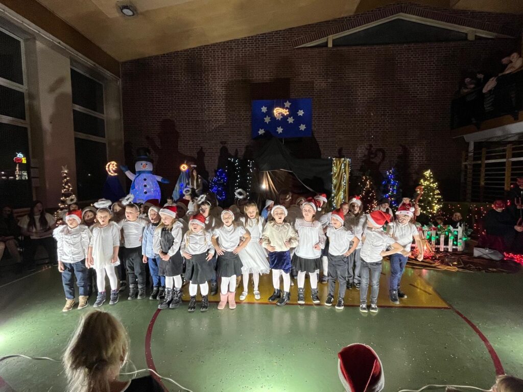 Na scenie w sali gimnastycznej stoi rząd najmłodszych uczniów klas pierwszych. Dzieci ubrane są w jasne stroje, wiele z nich ma na głowach czapki mikołajowe. Śpiewają lub recytują, stojąc obok siebie. W tle widoczna jest bożonarodzeniowa scenografia z choinkami, światełkami i dekoracją gwiazd.