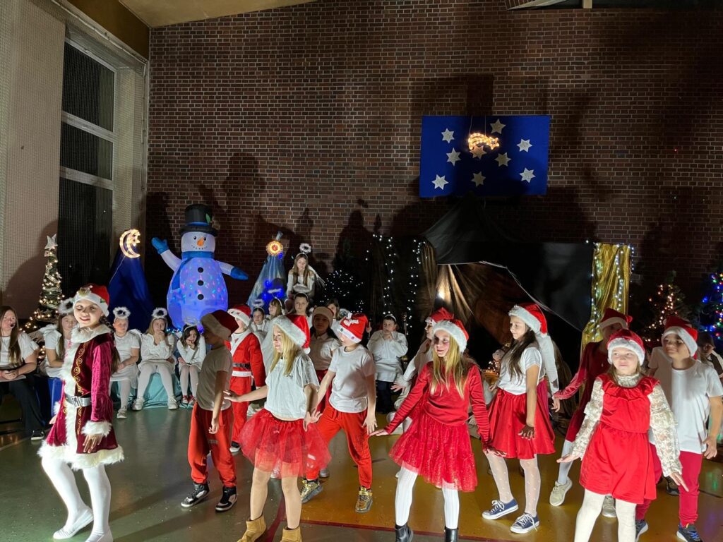 Grupa uczniów klas trzecich tańczy na scenie. Dzieci ubrane są w biało-czerwone stroje, część ma czapki mikołajowe. Stoją w kilku rzędach i wykonują ten sam układ taneczny. W tle znajdują się choinki, dekoracje świąteczne oraz inne dzieci obserwujące występ.