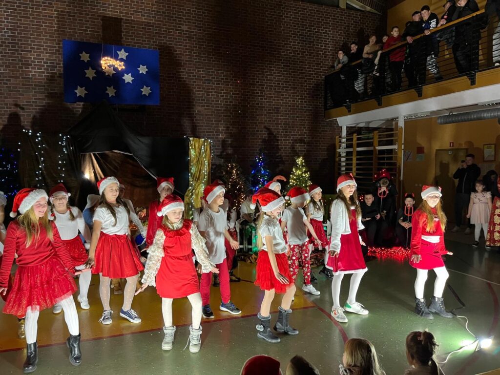 Uczniowie klas trzecich kontynuują taniec, stojąc w półokręgu na scenie. Dzieci mają na sobie czerwone spódnice, białe bluzki i czapki mikołajowe. Ich ręce są opuszczone lub lekko uniesione, zgodnie z choreografią. Widoczna jest publiczność oraz świąteczne dekoracje.