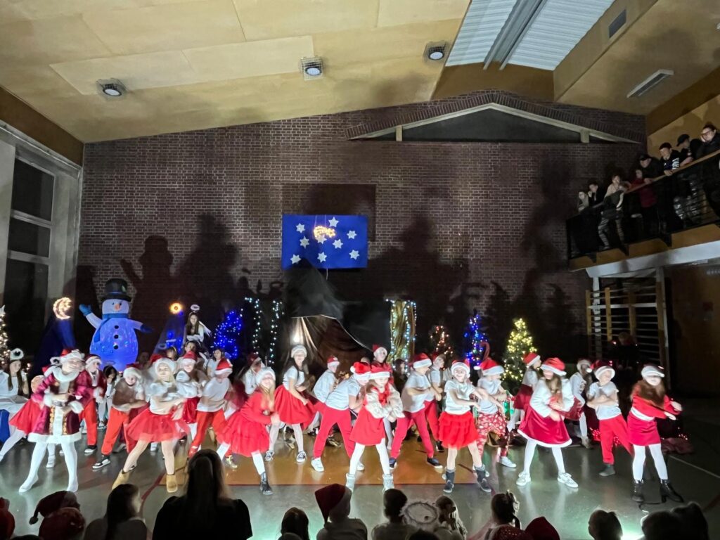 Duża grupa uczniów klas trzecich tańczy jednocześnie na całej szerokości sceny. Dzieci wykonują dynamiczny ruch, pochylając się i poruszając ramionami. W tle widać bogatą bożonarodzeniową scenografię z choinkami, światełkami i dekoracją gwiazd na ścianie.