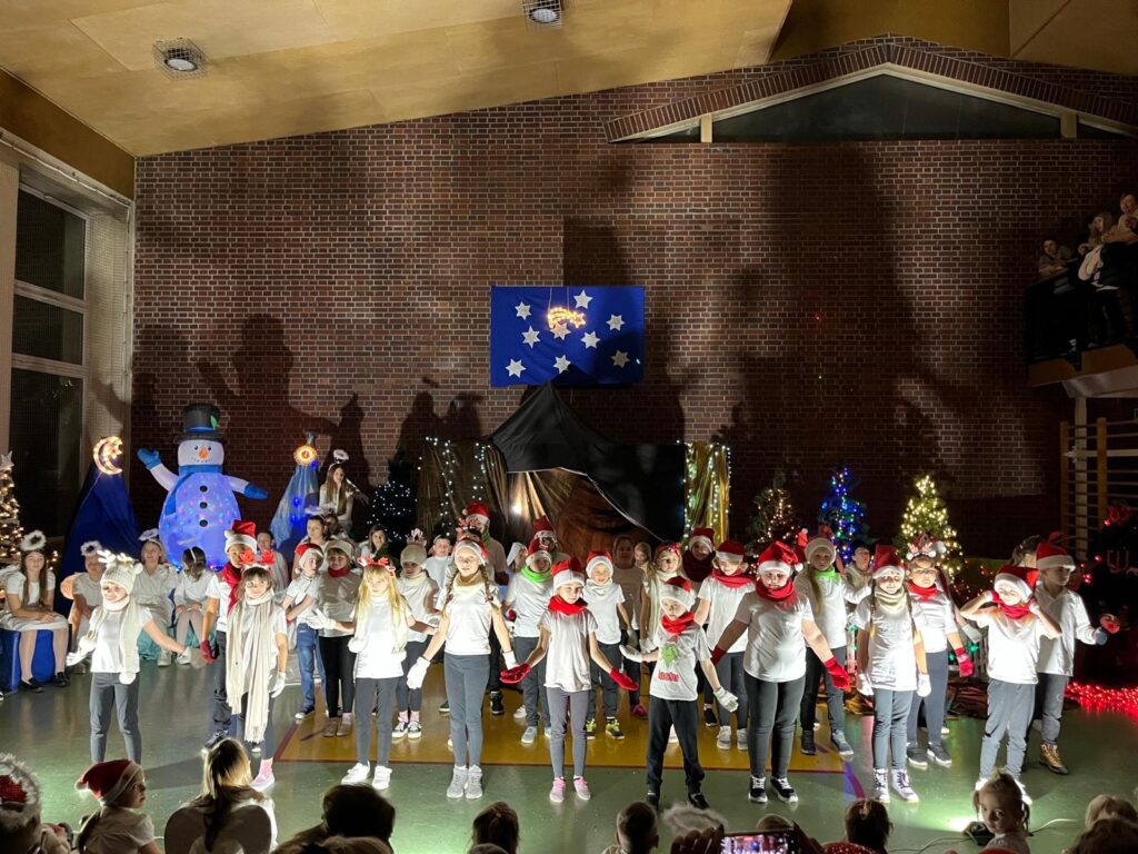 Na scenie stoi grupa uczniów klas drugich, ustawionych w kilku rzędach. Dzieci ubrane są w jasne koszulki, ciemne spodnie oraz kolorowe dodatki, m.in. czerwone szaliki i rękawiczki. Wszyscy wykonują wspólny układ taneczny, unosząc ręce do góry.