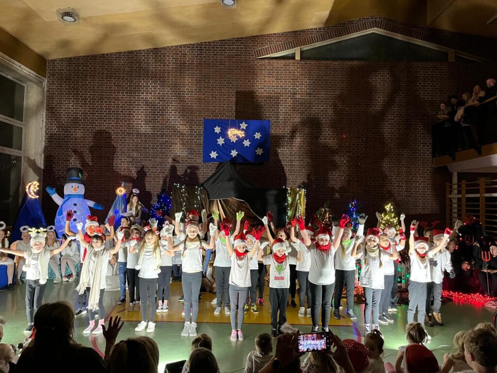 Uczniowie klas drugich stoją w szeregu na scenie z uniesionymi rękami, kończąc taniec. Dzieci są uśmiechnięte i zwrócone w stronę publiczności. Przed sceną widać zgromadzonych widzów, a w tle świąteczne dekoracje i choinki.