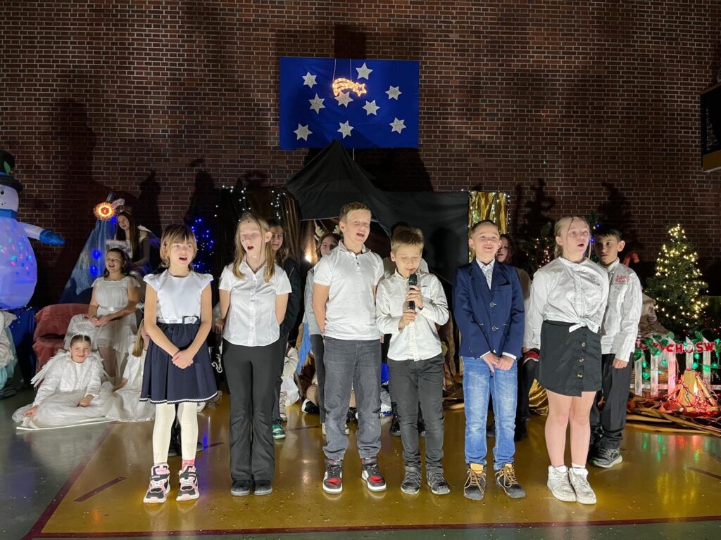 Na środku sceny stoi grupa uczniów klasy 4a. Dzieci ubrane są w eleganckie stroje: białe koszule, spódnice, spodnie oraz marynarki. Jedno z dzieci trzyma mikrofon i mówi lub śpiewa, a pozostali stoją obok, skupieni na występie. W tle widoczna jest stajenka i choinki.