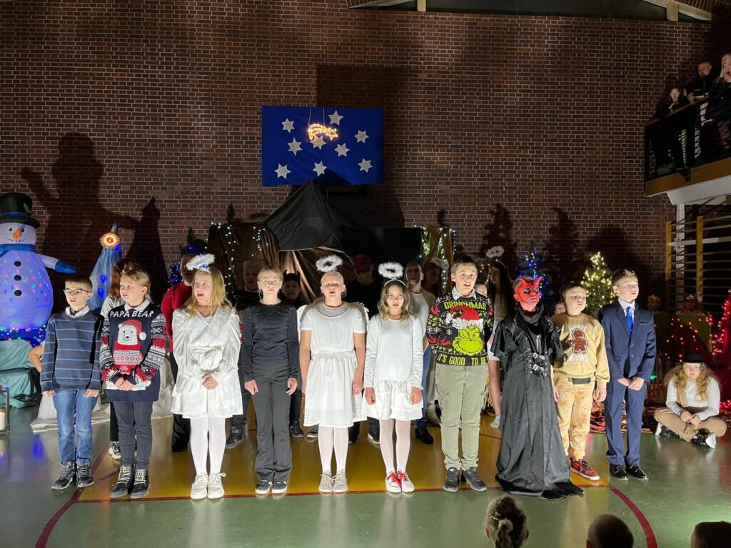 Uczniowie klasy 4b stoją w rzędzie na scenie. Część dzieci ma na sobie białe stroje, inne elementy ludowe lub świąteczne dodatki. Kilkoro uczniów trzyma mikrofony. Za nimi widoczne są anioły, dekoracje świąteczne i choinki.