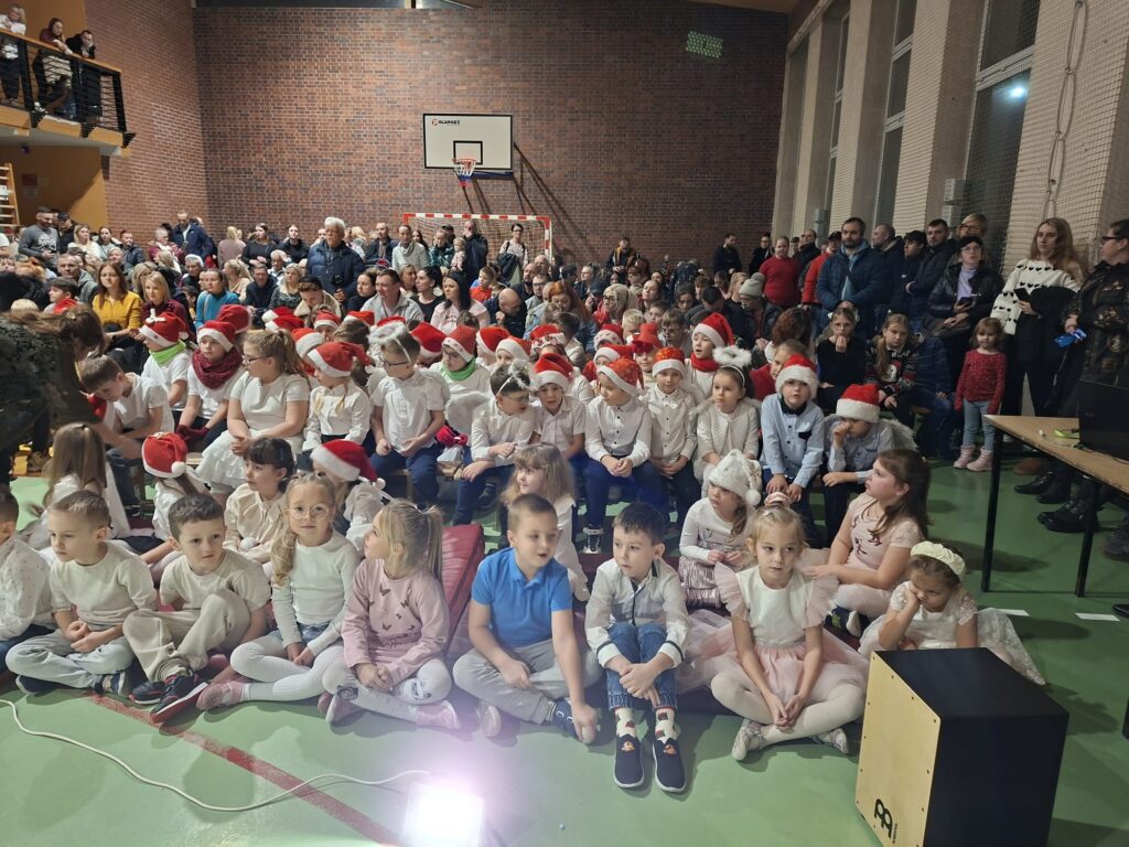 W sali gimnastycznej siedzi liczna publiczność złożona z dzieci, rodziców i nauczycieli. Na pierwszym planie na podłodze siedzą przedszkolaki w jasnych strojach i czapkach mikołajowych, a za nimi na trybunach i stojąc wokół – dorośli widzowie. Wszyscy zwróceni są w stronę sceny, oglądając występ.