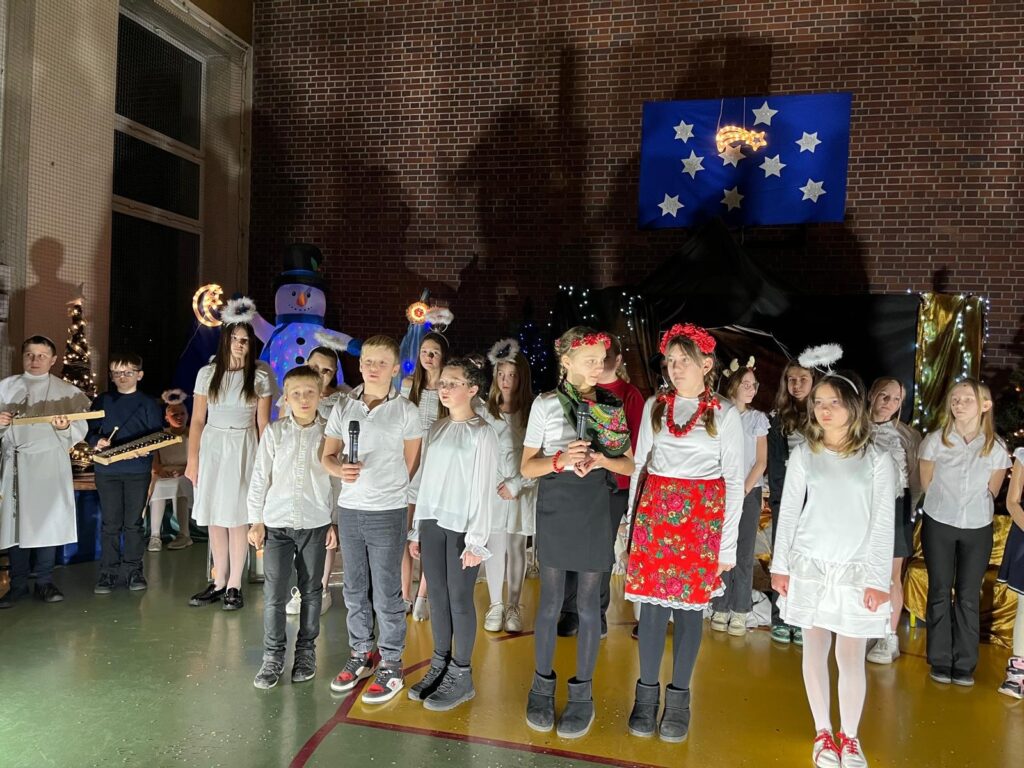 Na scenie stoi grupa uczniów tworzących szkolny zespół muzyczny. Dzieci trzymają mikrofony i instrumenty, a część śpiewa. Wszyscy zwróceni są w stronę publiczności. W tle widoczne są anioły, choinki oraz oświetlona bożonarodzeniowa scenografia.