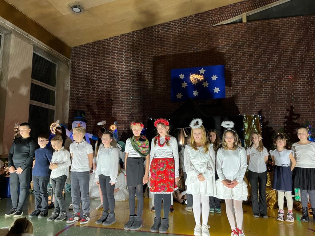 Szkolny zespół muzyczny stoi na scenie w pełnym składzie. Uczniowie śpiewają, trzymając mikrofony, a pozostali stoją obok w skupieniu. Scena jest bogato oświetlona, w tle widać dekorację gwiazd, stajenkę oraz choinki z lampkami.