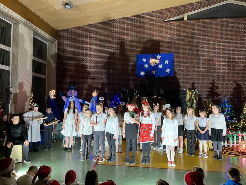 Na scenie stoi szkolny zespół muzyczny złożony z uczniów. Dzieci ustawione są w półokręgu i śpiewają, część z nich trzyma mikrofony. Po lewej stronie widoczni są uczniowie grający na instrumentach perkusyjnych i melodycznych. W tle znajdują się anioły, choinki oraz bożonarodzeniowa scenografia ze światłami.