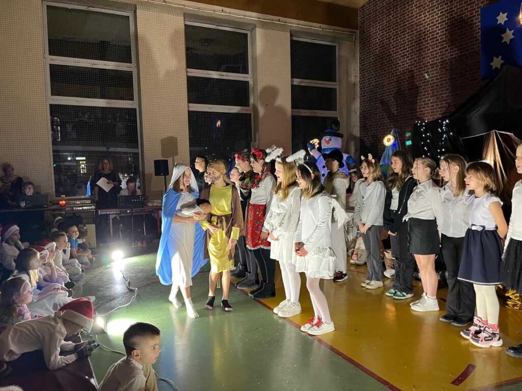 Na środku sceny uczennica przebrana za Maryję, ubrana w jasną suknię i niebieski płaszcz, trzyma na rękach lalkę symbolizującą Dzieciątko Jezus. Obok niej stoi uczeń w roli świętego Józefa. Wokół nich ustawieni są inni uczniowie przebrani za anioły i postacie jasełkowe. Na pierwszym planie, przy scenie, siedzą przedszkolaki oglądające przedstawienie, a całość dopełnia oświetlona bożonarodzeniowa dekoracja.