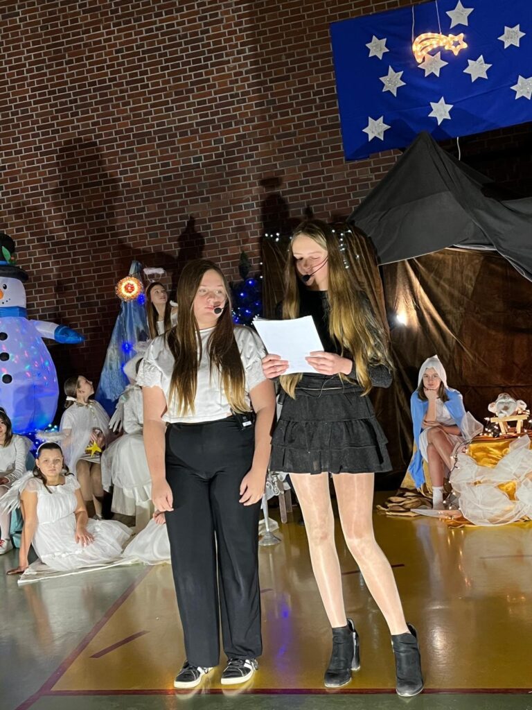 Dwie uczennice stoją na środku sali, trzymając kartki z tekstem i mikrofony nagłowne. Jedna ubrana jest w czarną spódnicę i bluzkę, druga w jasną bluzkę i ciemne spodnie. Czytają tekst narracji. W tle widoczne są dzieci przebrane za anioły oraz elementy bożonarodzeniowej dekoracji.