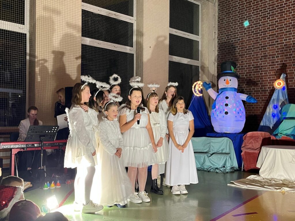 Grupa dziewczynek ubranych w białe sukienki stoi razem na scenie. Mają na głowach opaski z aureolami, a jedna z nich trzyma mikrofon i śpiewa lub recytuje. Pozostałe dzieci stoją obok, skupione i zwrócone w stronę publiczności. W tle widoczne są choinki, światełka i bożonarodzeniowa scenografia.