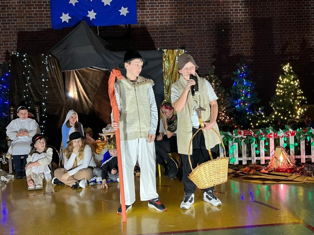 Dwóch chłopców stoi na środku sceny, przebranych za pasterzy. Mają na sobie jasne koszule, kamizelki i nakrycia głowy. Jeden trzyma długi kij pasterski, drugi kosz wiklinowy i mikrofon. Za nimi siedzą inne dzieci w kostiumach, a całość otacza świąteczna dekoracja z choinkami i oświetleniem.