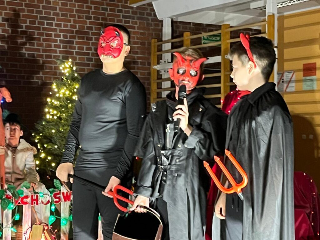 Trzech chłopców ubranych w czarne stroje stoi obok siebie. Mają czerwone maski diabłów oraz trójzęby trzymane w dłoniach. Jeden z nich mówi do mikrofonu. W tle widać świąteczną scenografię i inne dzieci obserwujące występ.