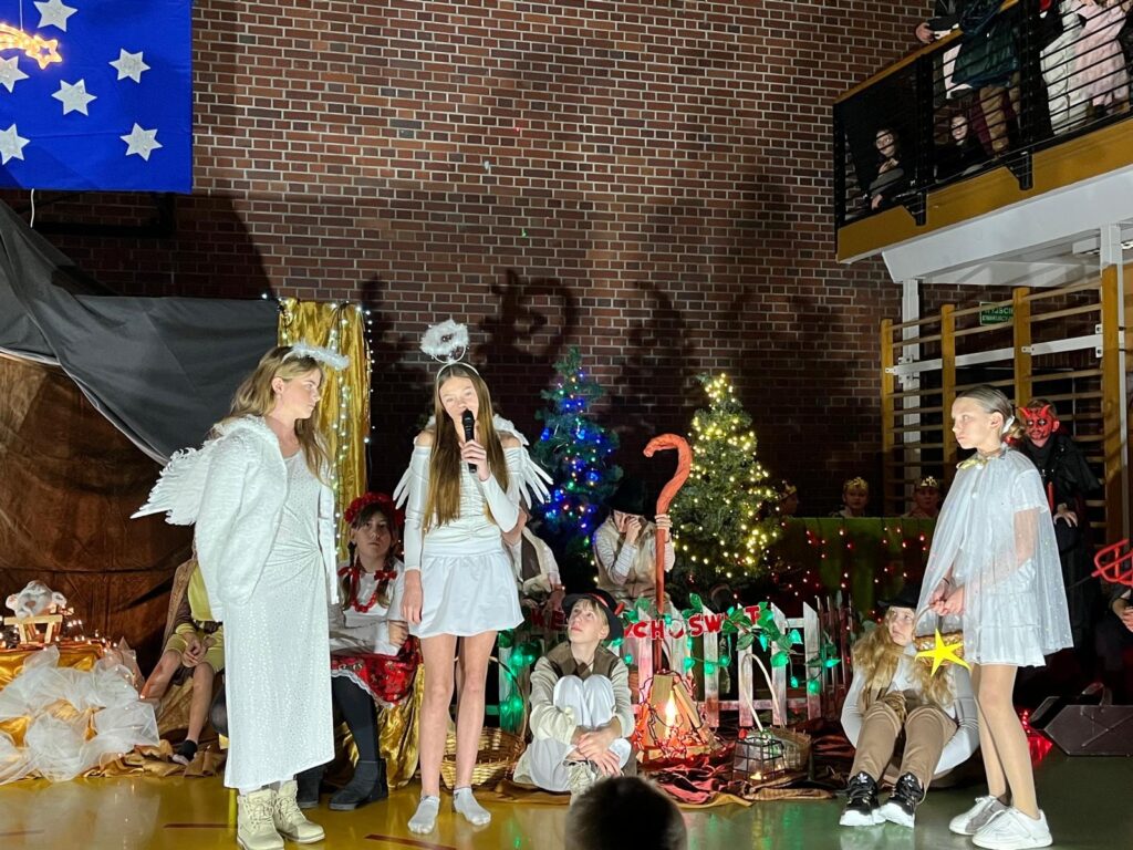 Na scenie znajduje się grupa dzieci w różnorodnych kostiumach jasełkowych – aniołów, pasterzy i postaci ludowych. Jedna z dziewczynek z aureolą trzyma mikrofon i mówi, a pozostałe dzieci siedzą lub stoją wokół niej, słuchając. W tle widać choinki, światełka i dekoracje symbolizujące stajenkę.