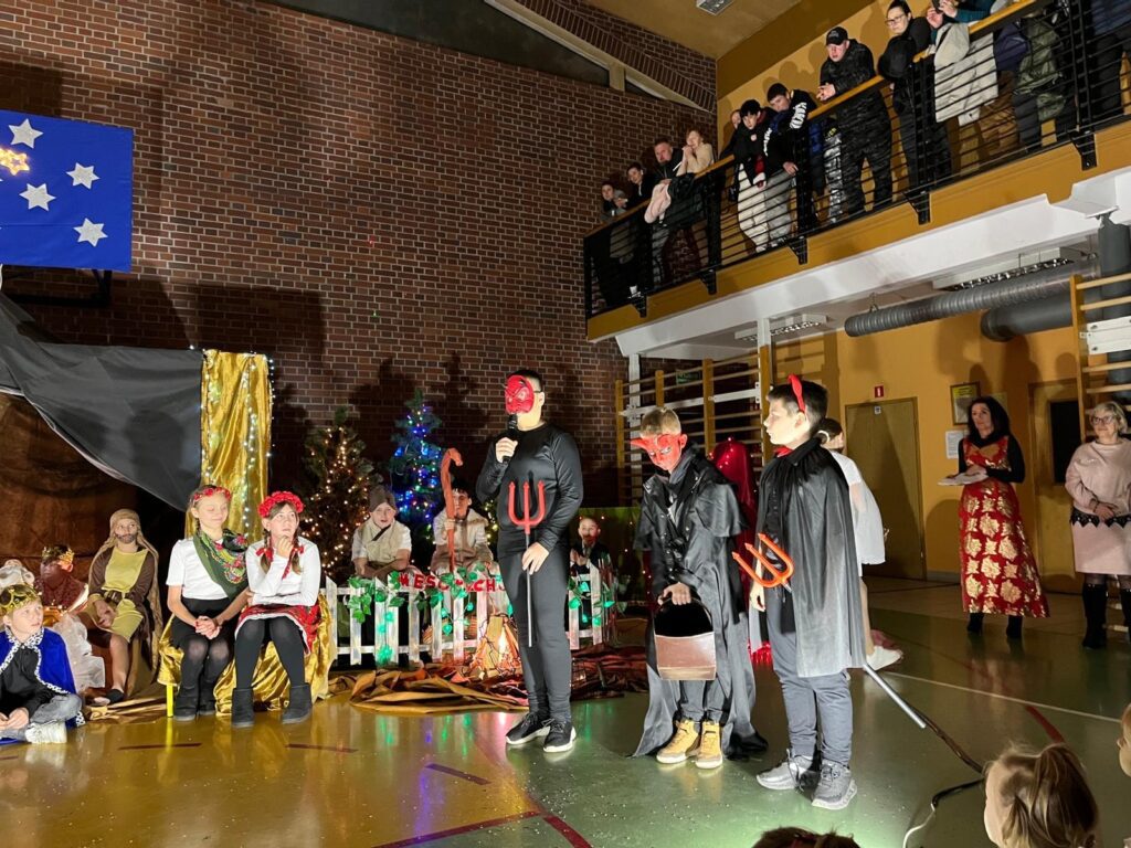 Na środku sceny stoją uczniowie przebrani za diabły, ubrani na czarno i wyposażeni w czerwone maski oraz trójzęby. Jeden z nich przemawia do mikrofonu, pozostali stoją obok. Za nimi widoczne są dzieci w innych rolach oraz świąteczna dekoracja z choinkami i światłami.