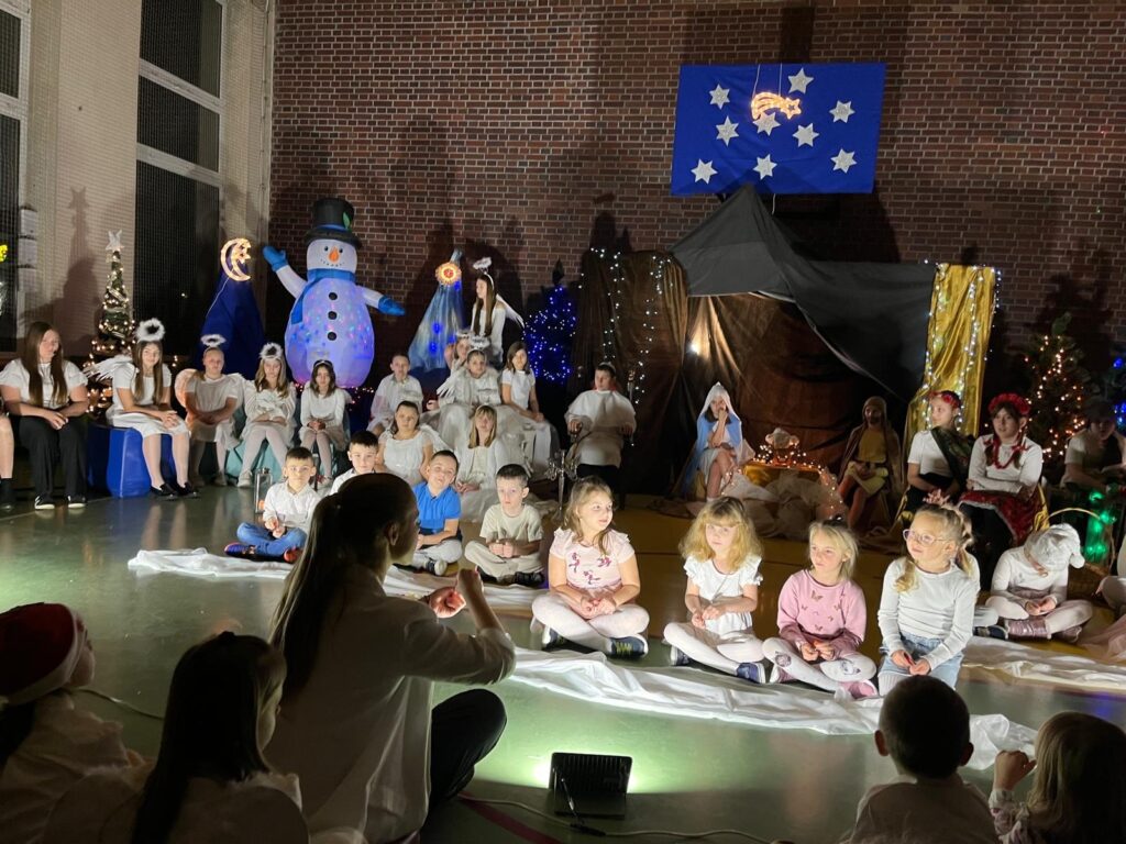 Na pierwszym planie stoi rząd małych dzieci w wieku przedszkolnym, ubranych w jasne stroje. Dzieci trzymają w dłoniach małe instrumenty muzyczne i śpiewają. Przed nimi ustawione są kolorowe dzwonki. W tle widać starsze dzieci oraz świąteczną scenografię.