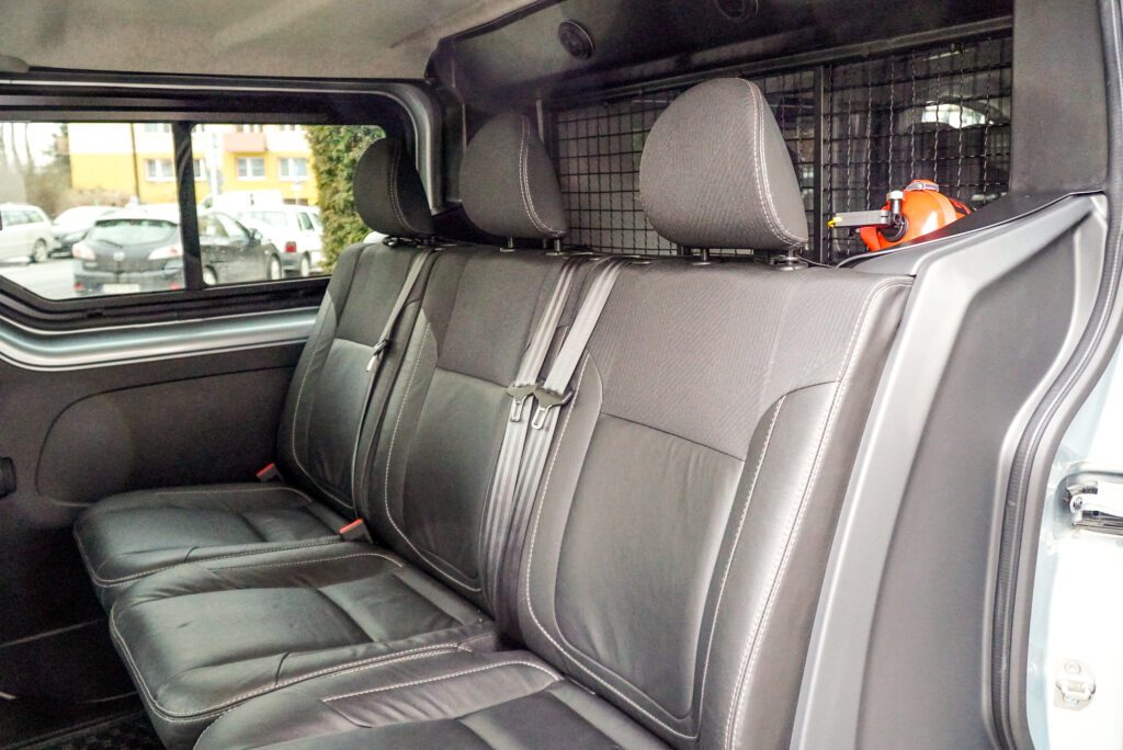 Wnętrze samochodu, widok na tylny rząd trzech czarnych foteli. Za oparciami znajduje się czarna, metalowa krata oddzielająca bagażnik. Po prawej stronie, za kratą, widoczna cześć pomarańczowej gaśnicy.