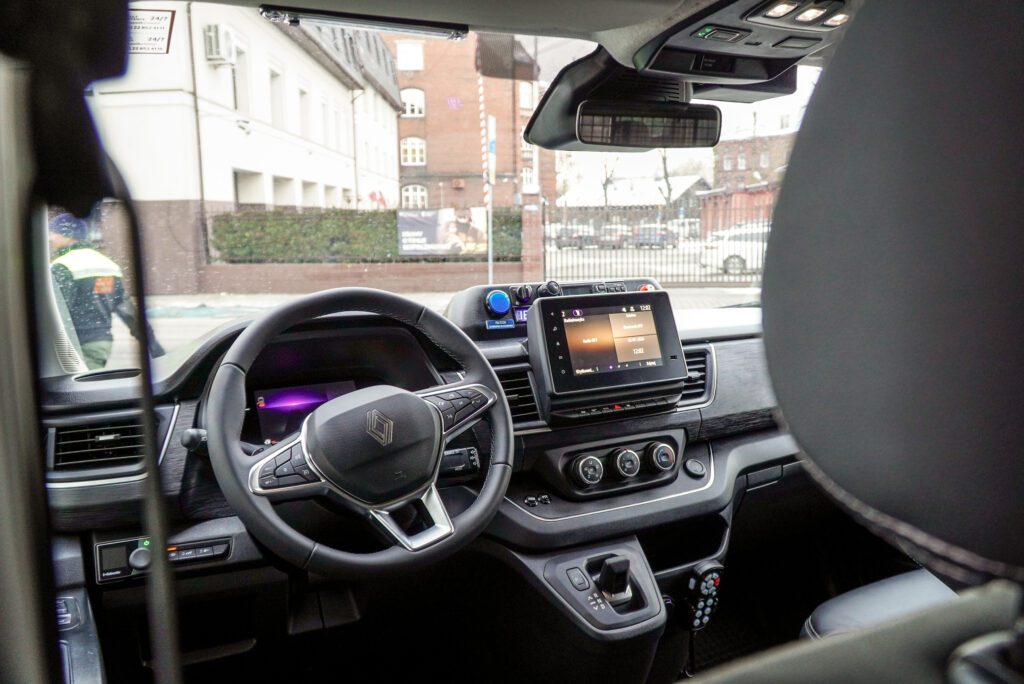 Wnętrze samochodu widziane z perspektywy tylnej kanapy. Na pierwszym planie ciemna kierownica z logo Renault, deska rozdzielcza z ekranem dotykowym i drążek automatycznej skrzyni biegów. Przez przednią szybę widać budynek i ogrodzenie.