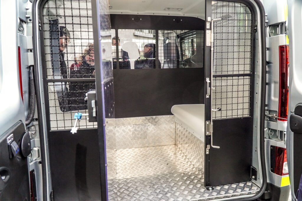 Widok do wnętrza tylnej części furgonetki przez otwarte drzwi. Pomieszczenie jest oddzielone czarną kratą. W środku widać jasną ławkę, metalową podłogę oraz ścianę grodziową z małym zakratowanym okienkiem.